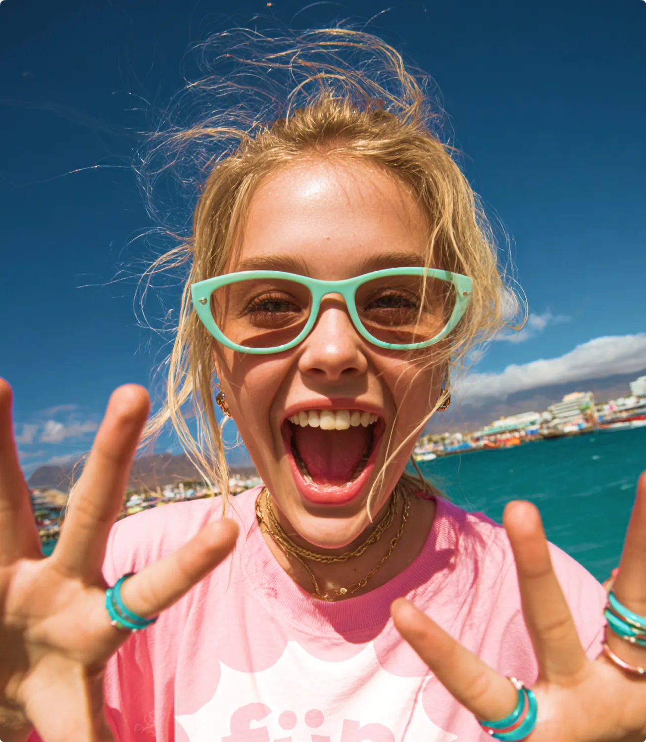 Cheering blonde girl in mint sunglasses and a pink aqua fun t-shirt throwing up her hands in front of a marina and turquoise sea