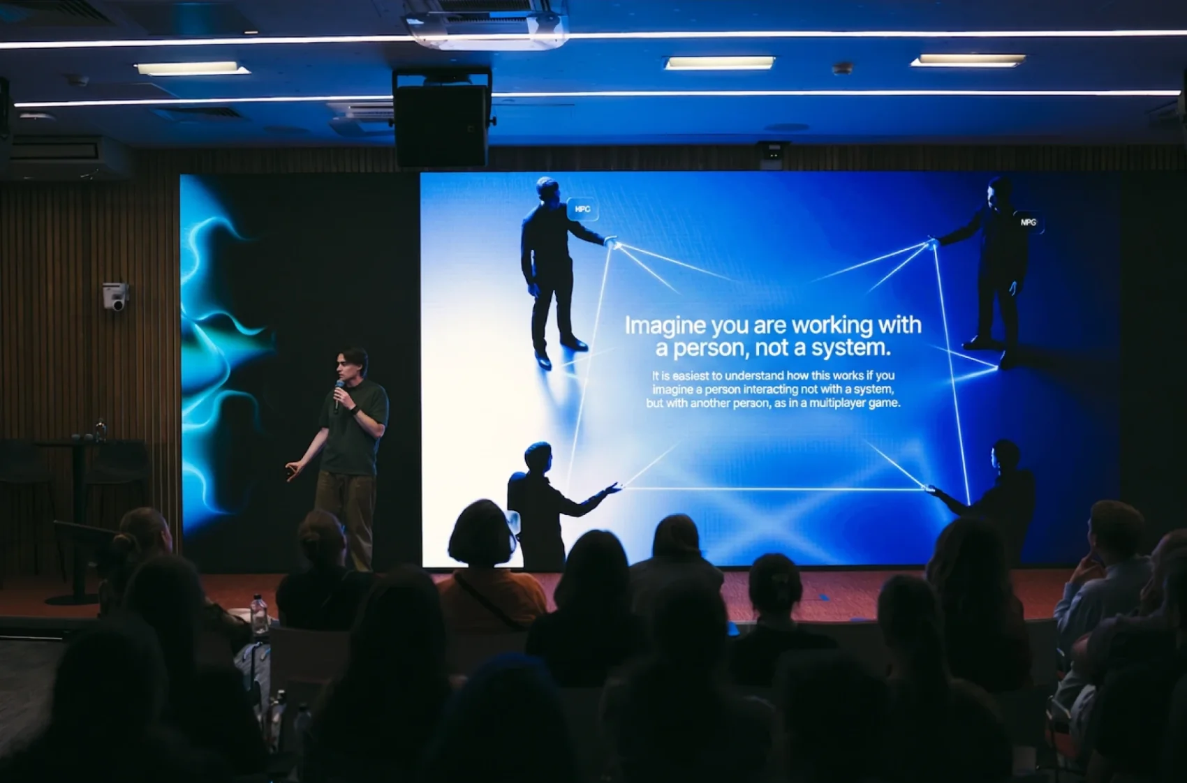 A conference talk on designing AI features, the slide behind the speaker reading "Imagine you are working with a person, not a system"